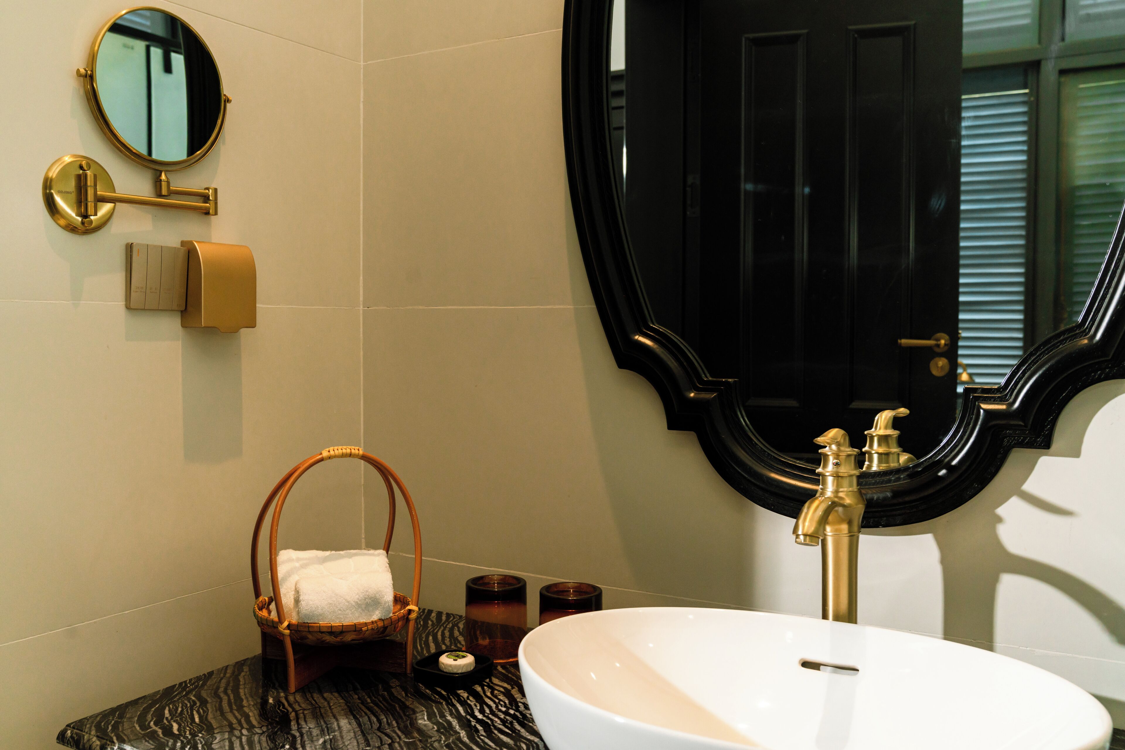 Een badkamer met een grote ronde spiegel, een wit wastafelmeubel, een gouden kraan, een zwarte deur en een klein rieten mandje met een witte handdoek.