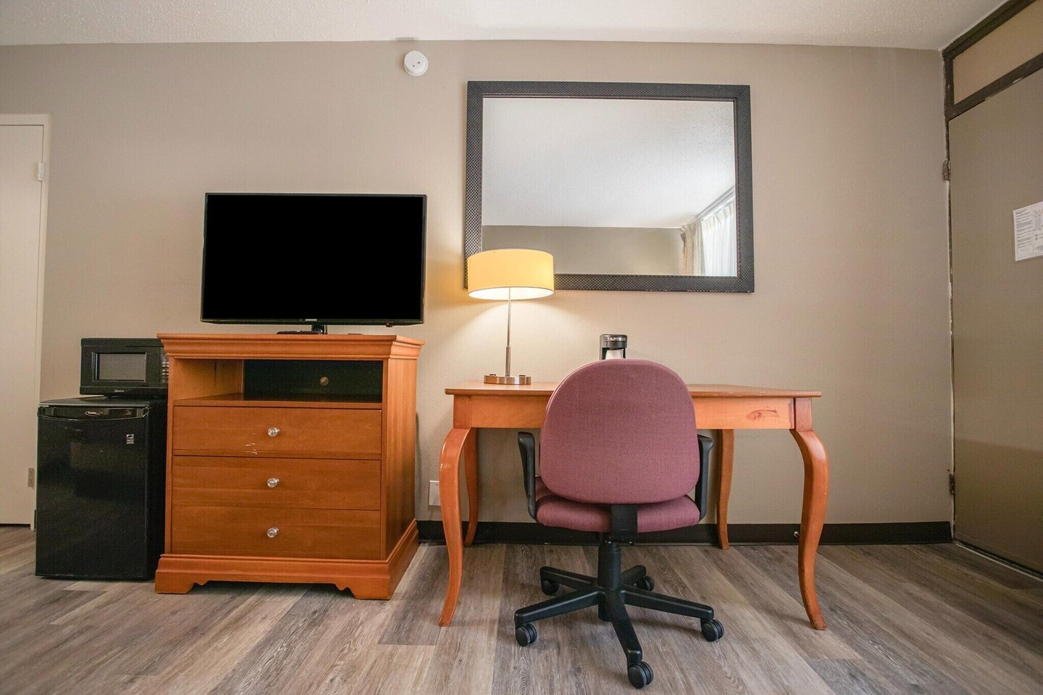 A hotel room with a wooden desk, a television, a lamp, a chair, a microwave, and a refrigerator.