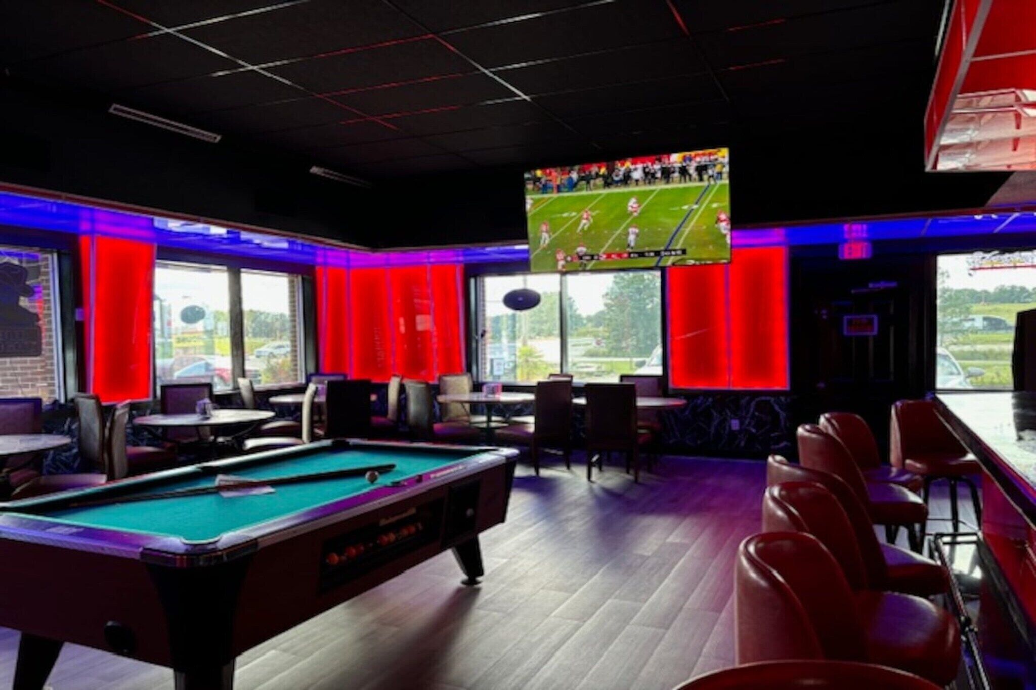 Une table de billard dans une pièce éclairée de lumières rouges, un grand écran diffusant un match sportif et une vue sur l’extérieur grâce à de grandes fenêtres.