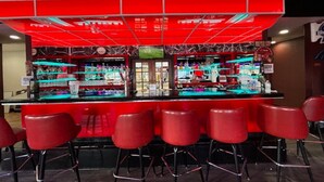 Zona de bar amb un mostrador vermell, diverses banquettes i prestatges plens de botelles.