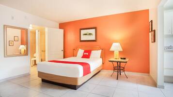 A hotel room with a bed, a bedside table with a lamp, a framed picture on the wall, and a door leading to another room.