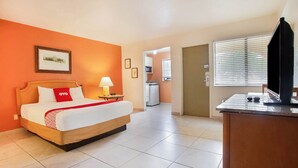 Habitación de hotel con cama, escritorio, lámpara, un cuadro en la pared y una cocina al fondo.