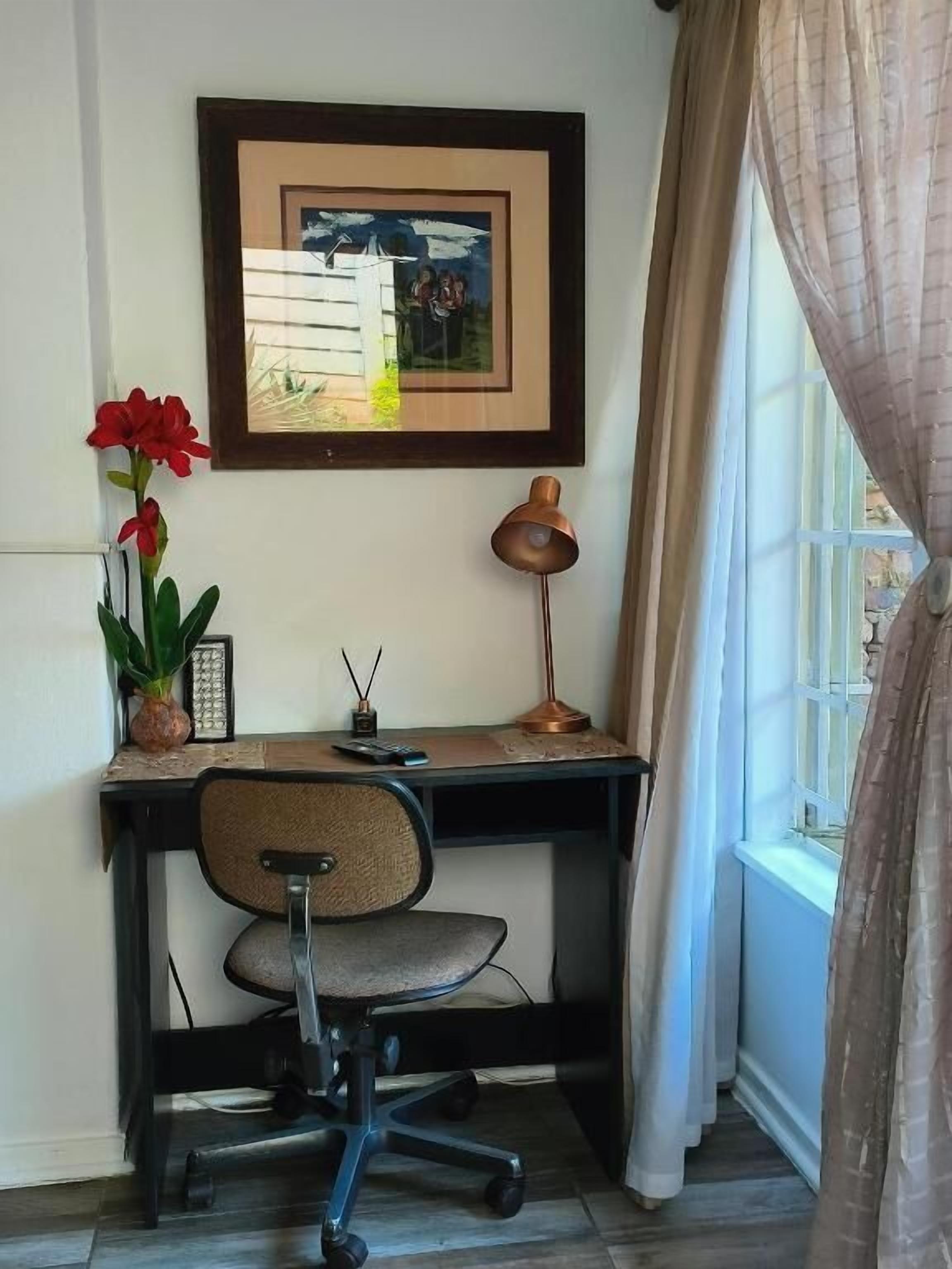 A corner office with a desk, chair, framed artwork, a potted plant, and a window with curtains.