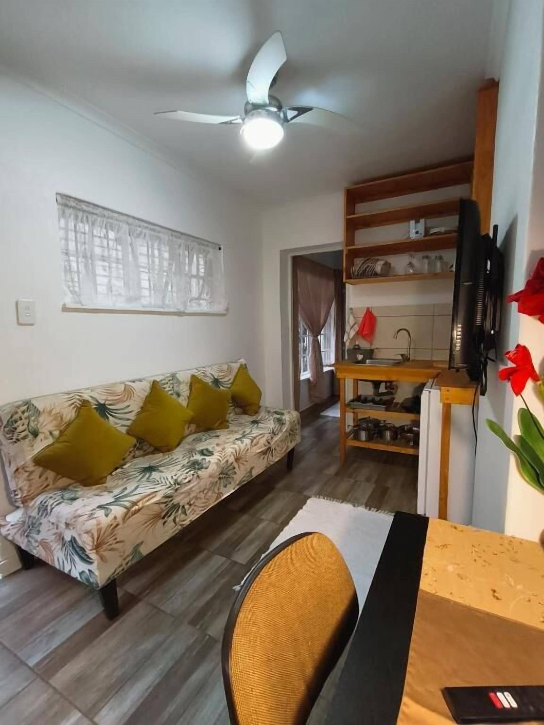 Una sala de estar con sofá, mesa de comedor, ventilador de techo y una zona de cocina con estantes y televisor.