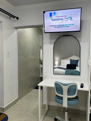 A hotel room with a bed, a desk with a chair, a television mounted on the wall, and a mirror above the desk.