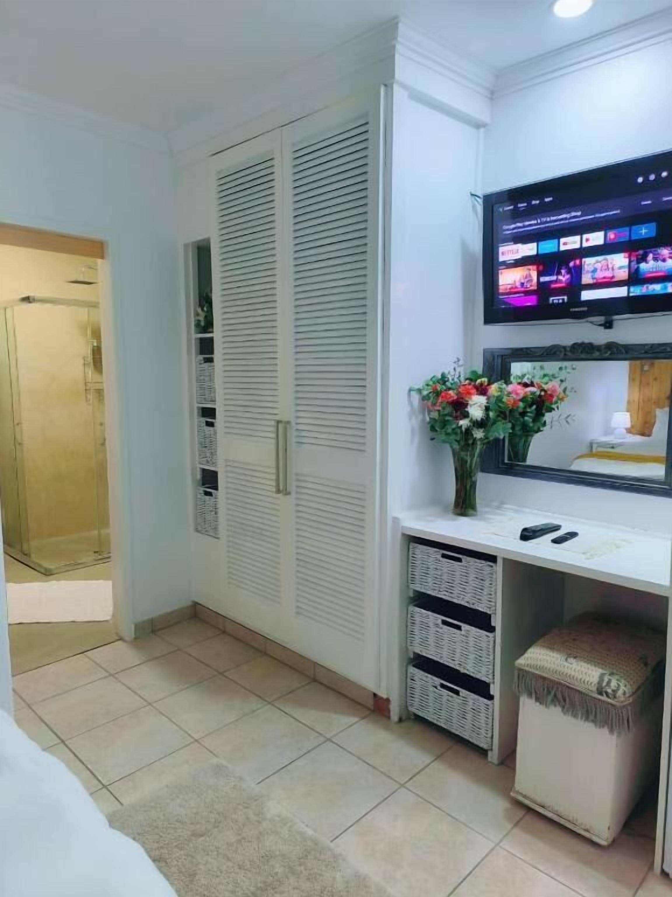 Une pièce avec un téléviseur fixé au mur, un bureau blanc orné d’un vase de fleurs et une armoire aux portes coulissantes.