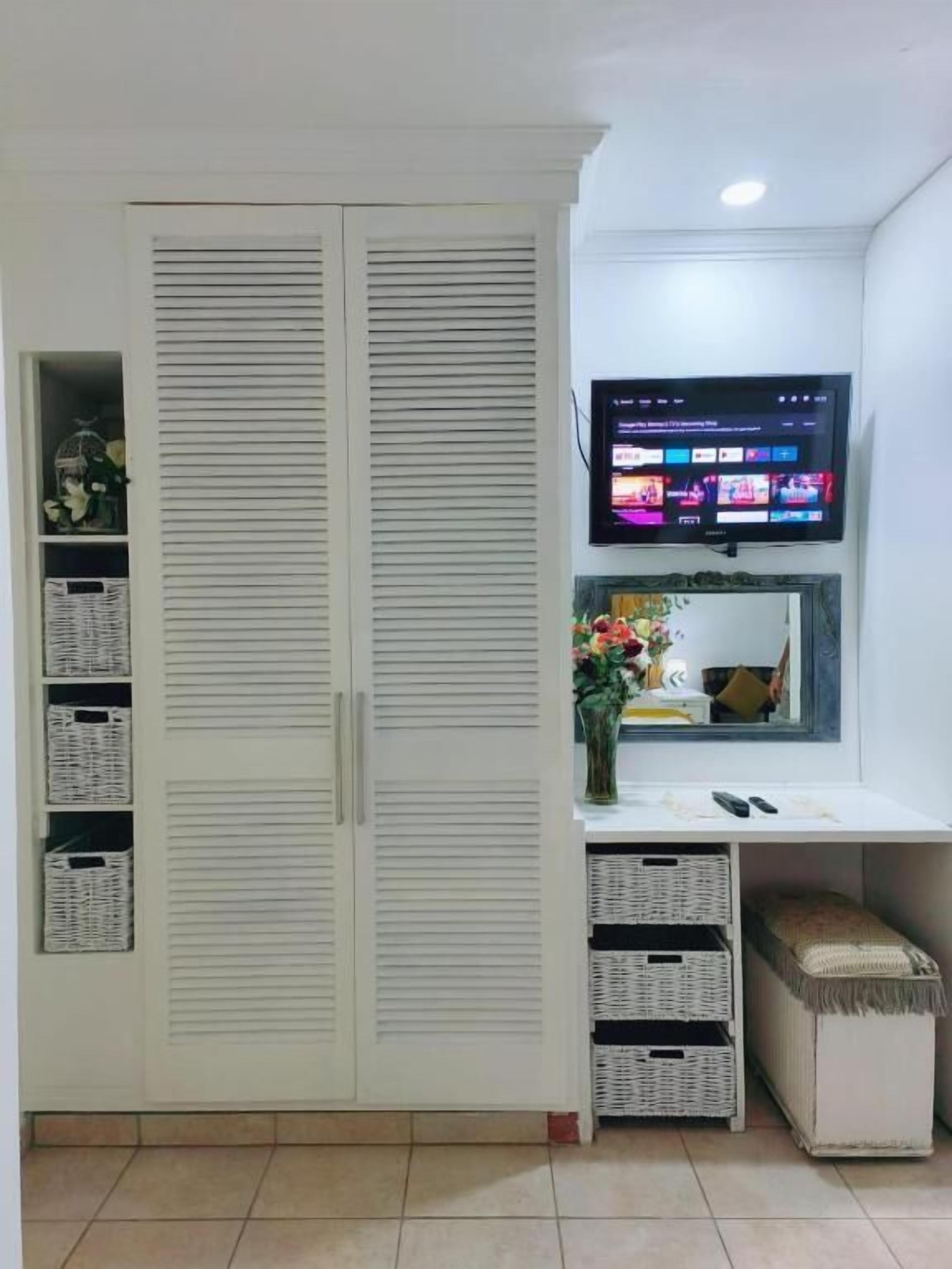 Une pièce avec une armoire blanche, un téléviseur à écran plat fixé au-dessus d’un bureau et des paniers de rangement.