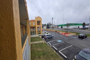 Un parking rempli de voitures, un bâtiment à la façade jaune et, au fond, une structure surmontée d’un toit végétalisé.