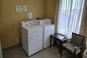 A laundry room with two washers and dryers, a small table, a chair, and a window with sheer curtains.