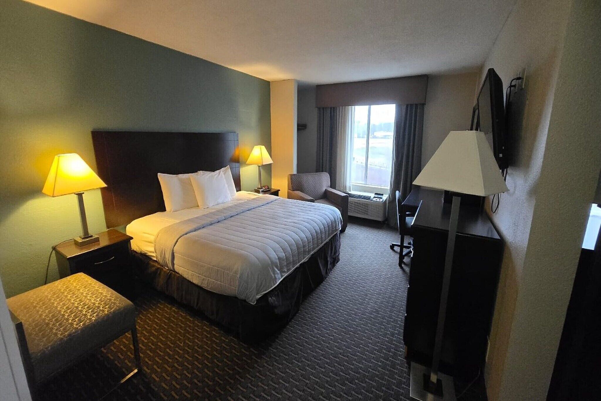 Quarto de hotel com cama, duas luminárias de cabeceira, escrivaninha, cadeira, televisão e janela com cortinas.