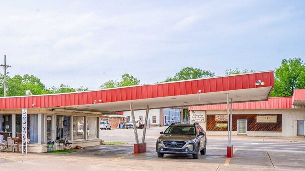 Una estación de servicio con toldo rojo, un coche aparcado debajo y una pequeña zona de estar al aire libre.