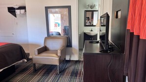 A hotel room with a bed, a chair, a desk with a computer, a mirror, and a bathroom with a sink and mirror.