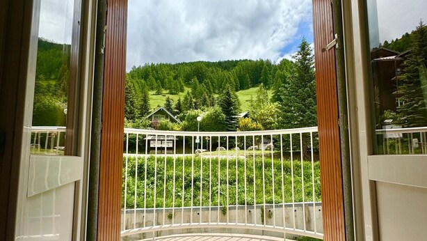 A room with a large glass door leading to a balcony with a view of a green landscape and a wooden fence. - STUDIO LA FOUX D'ALLOS (Allos)