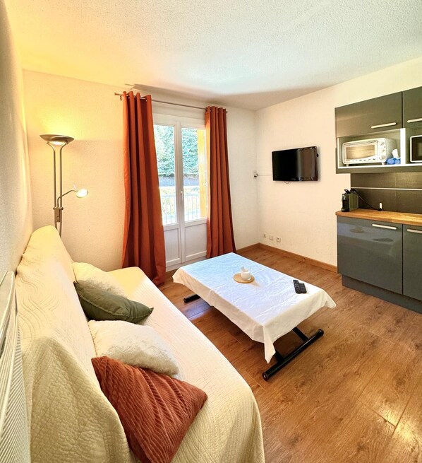 A compact room with a sofa, a small table, a TV, and a kitchenette.