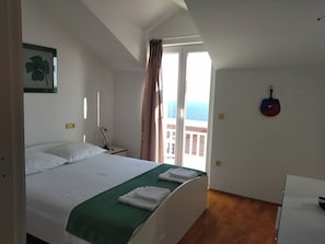 A bedroom with a bed, a green blanket, a white pillow, a desk, a chair, a guitar on the wall, and a balcony with a view. - Apartman Nada (Cavtat)