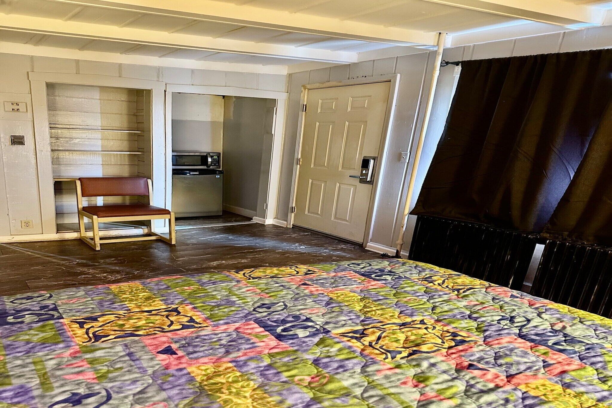A room with a bed, a chair, a door, and a small kitchenette.