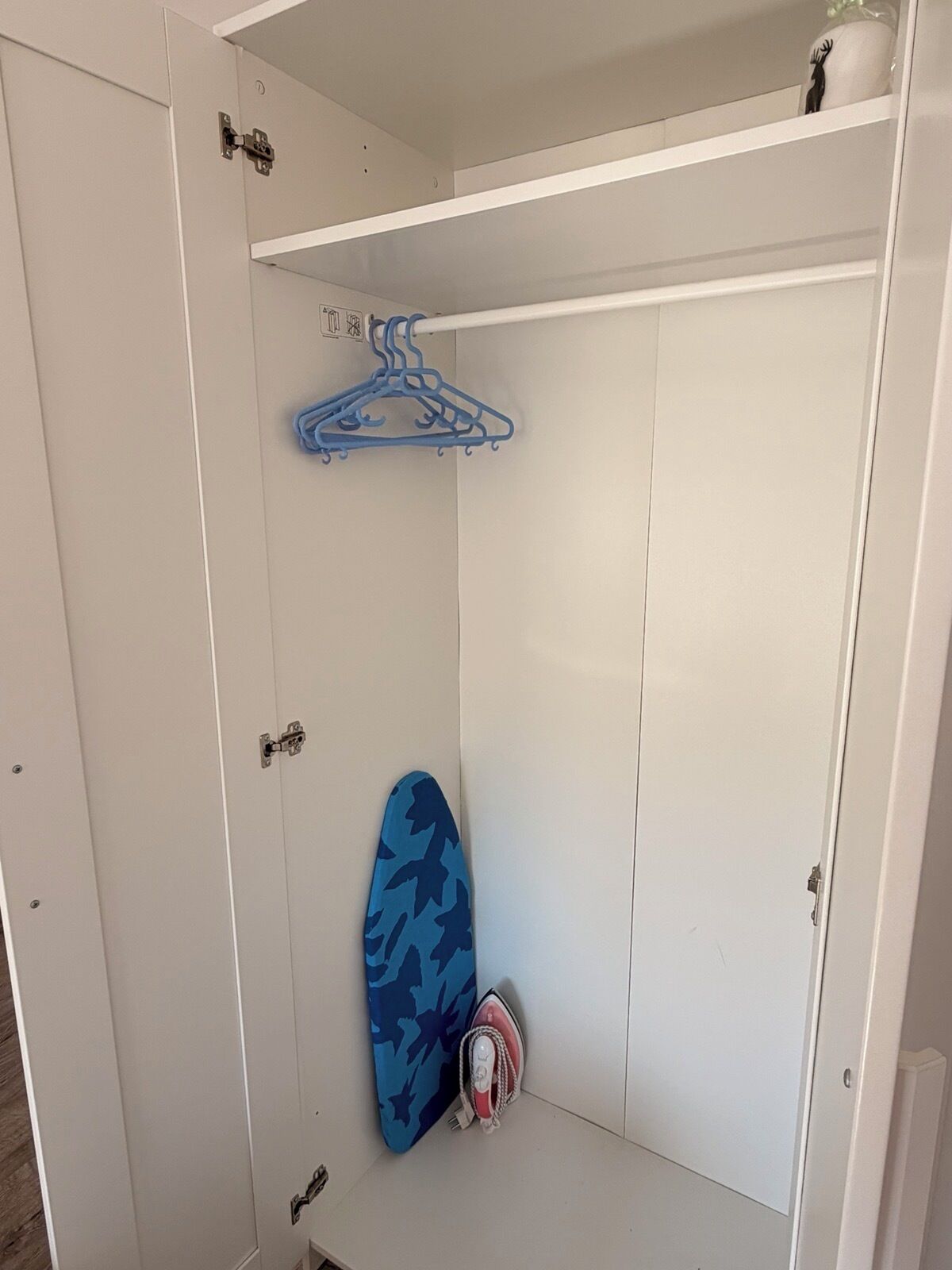 Ein weißer Kleiderschrank mit Kleiderstange, einem Surfbrett und einem Bügeleisen.