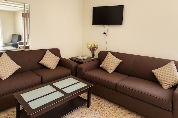 A living room with two brown sofas, a coffee table, a flat-screen TV, a small side table with a vase of flowers, and a mirror.