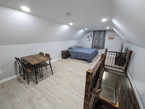 Una stanza con un tavolo da pranzo in legno, un letto con coperta blu, un tavolino con una sedia e una scala con corrimano in legno.