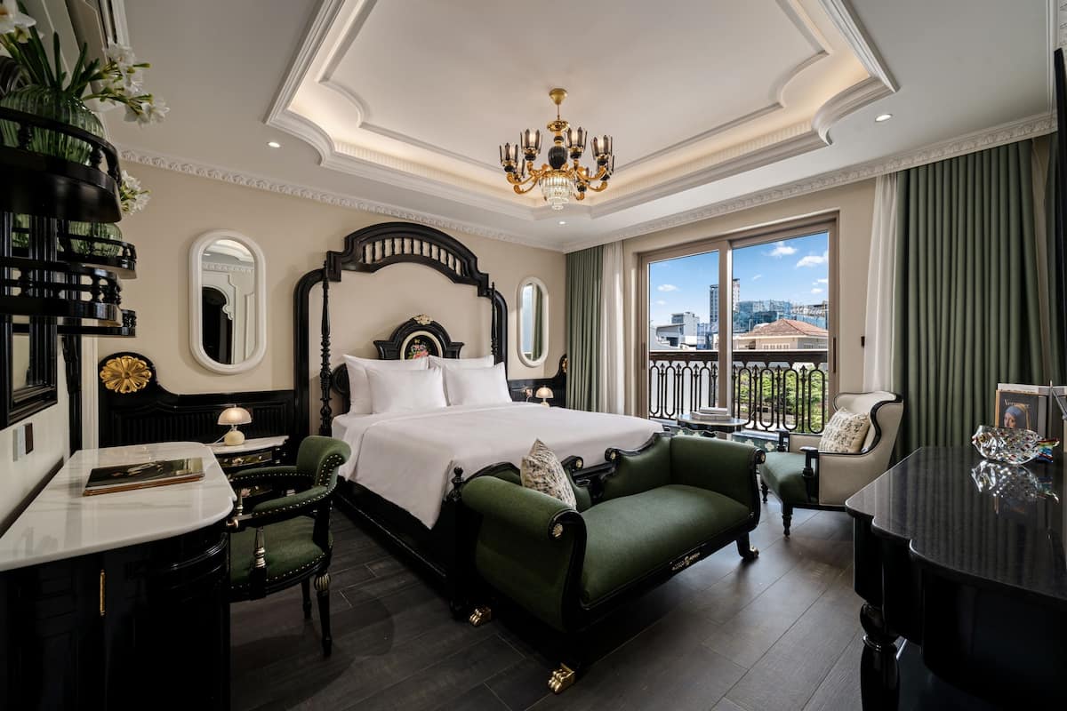 Quarto de hotel com uma cama grande, um espaço de estar com sofá verde, uma mesa com luminária e vista para a cidade através de grandes janelas.