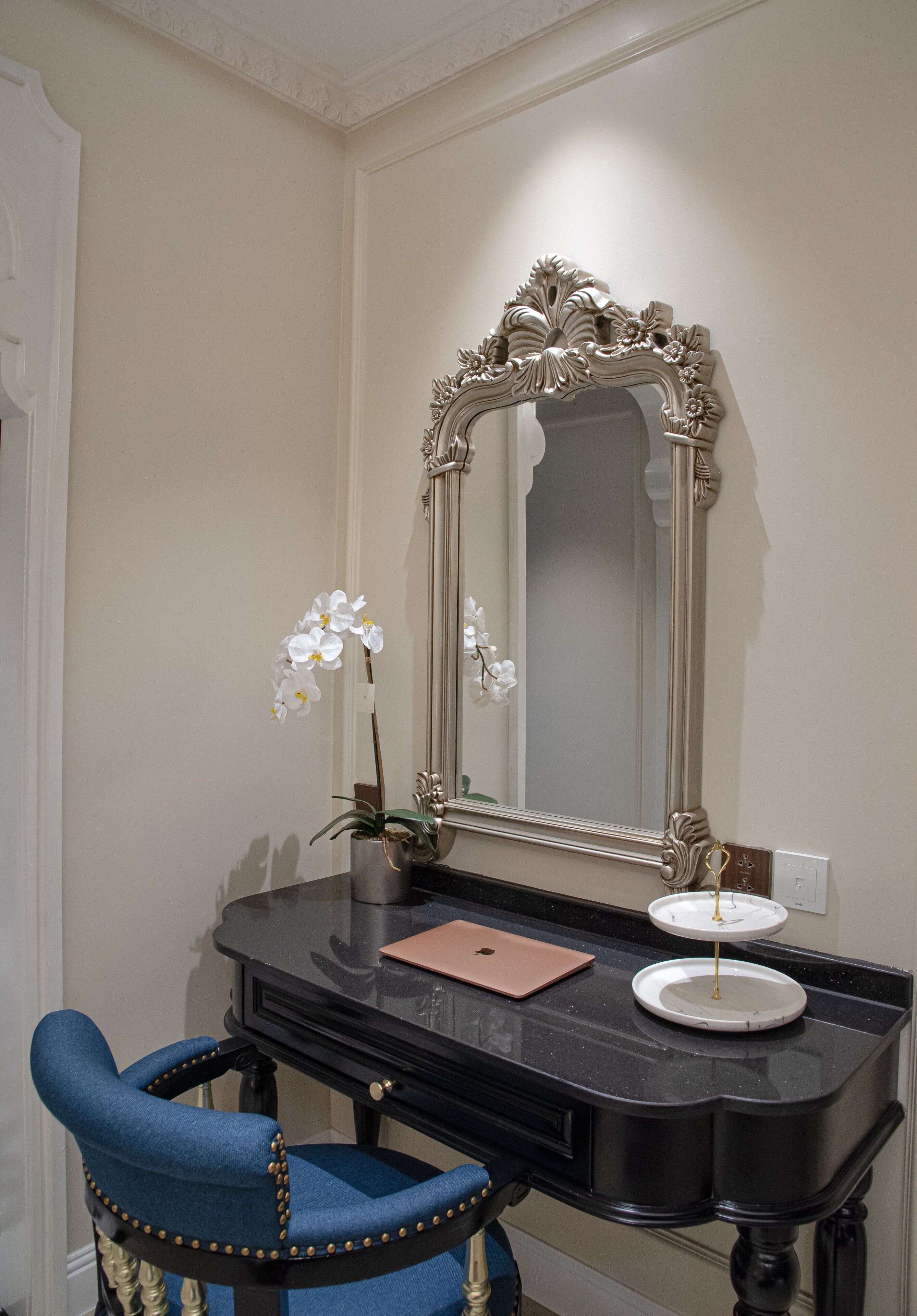 Une coiffeuse noire avec un grand miroir, une chaise bleue, une petite orchidée en pot et un ordinateur portable posé sur le comptoir.