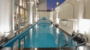 En indendørs swimmingpool med klare blå fliser, omgivet af dekorative hvide søjler og et højt loft.