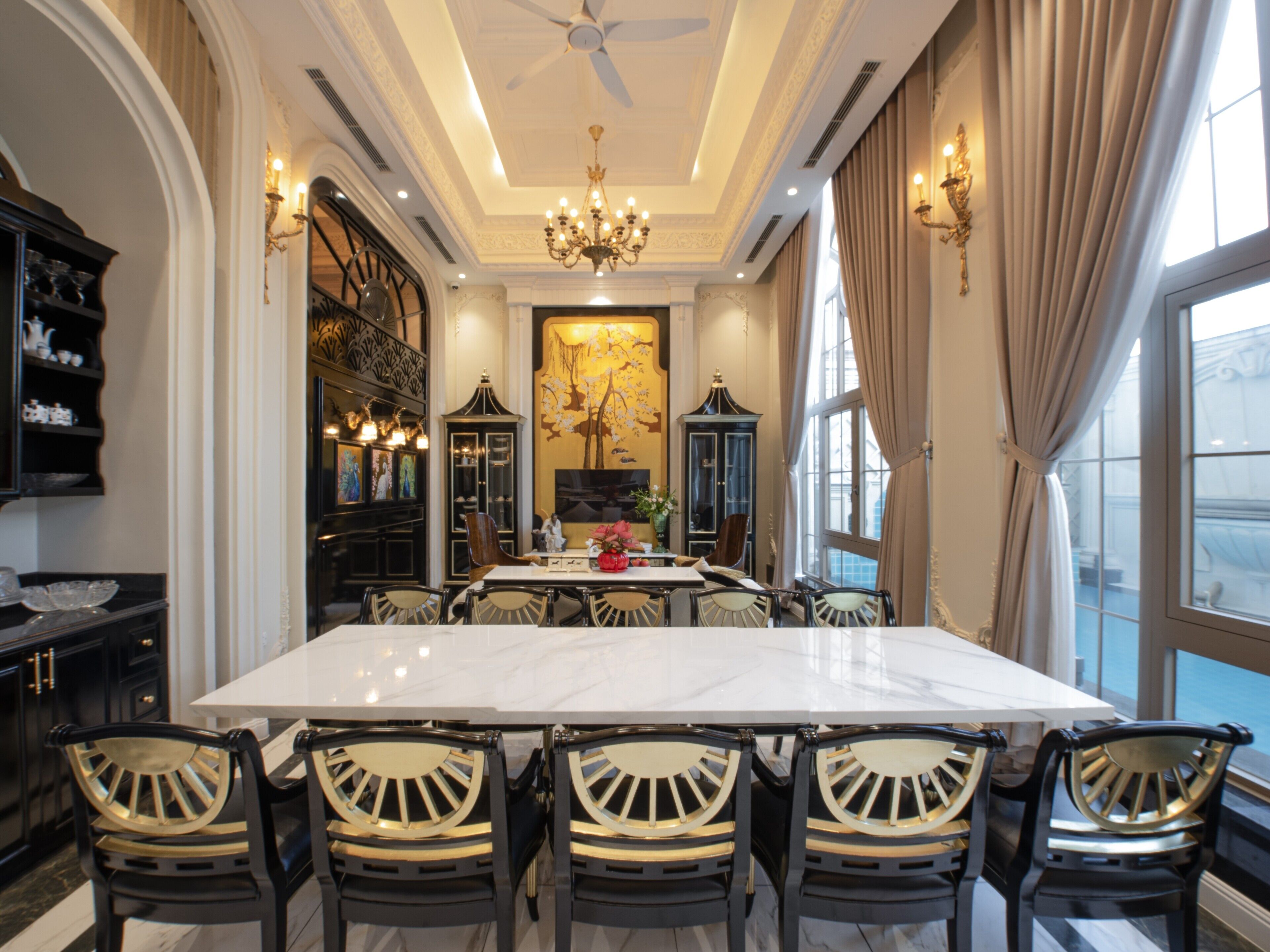 A dining room with a large marble table, black chairs with gold accents, a chandelier, and a large window with curtains.