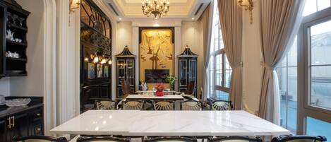 A dining room with a large marble table, black chairs with gold accents, a chandelier, and a large window with curtains.