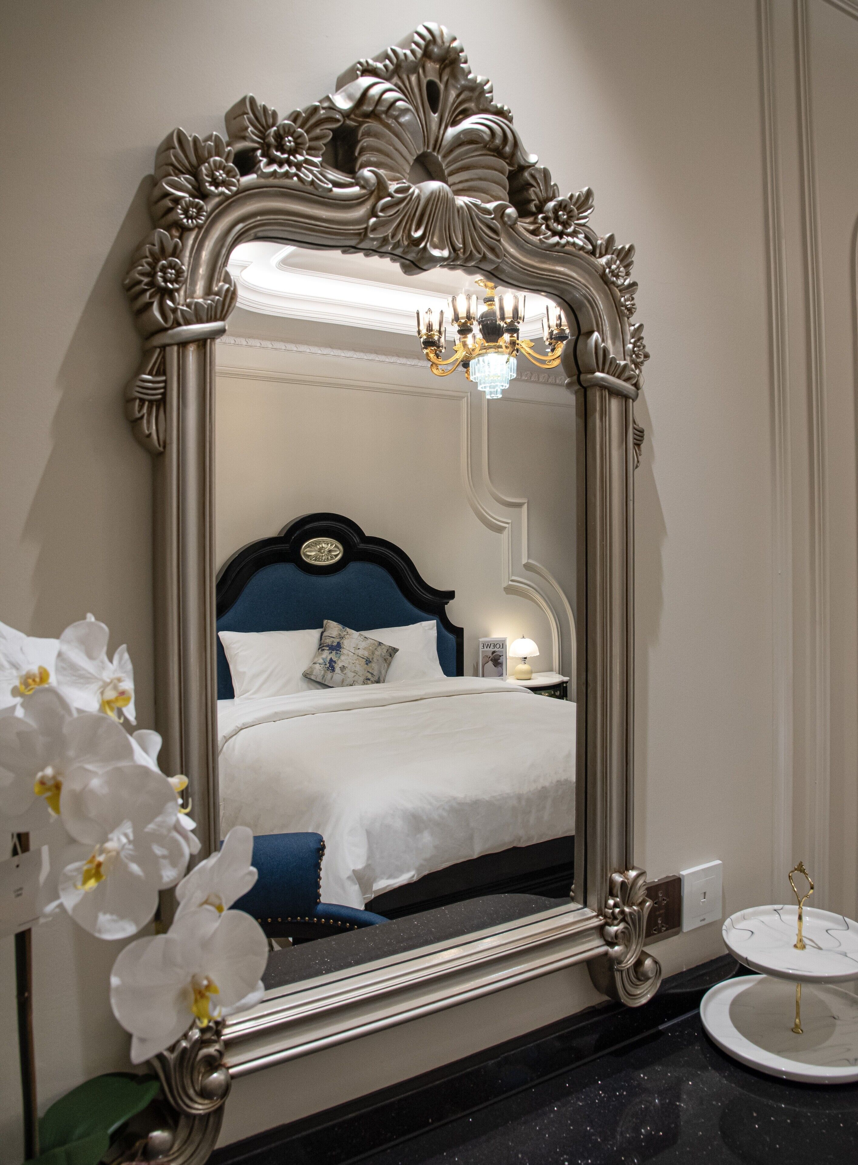 A mirror with an ornate frame, a bed with a blue headboard, a bedside table with a lamp, and a small round table with a decorative item.