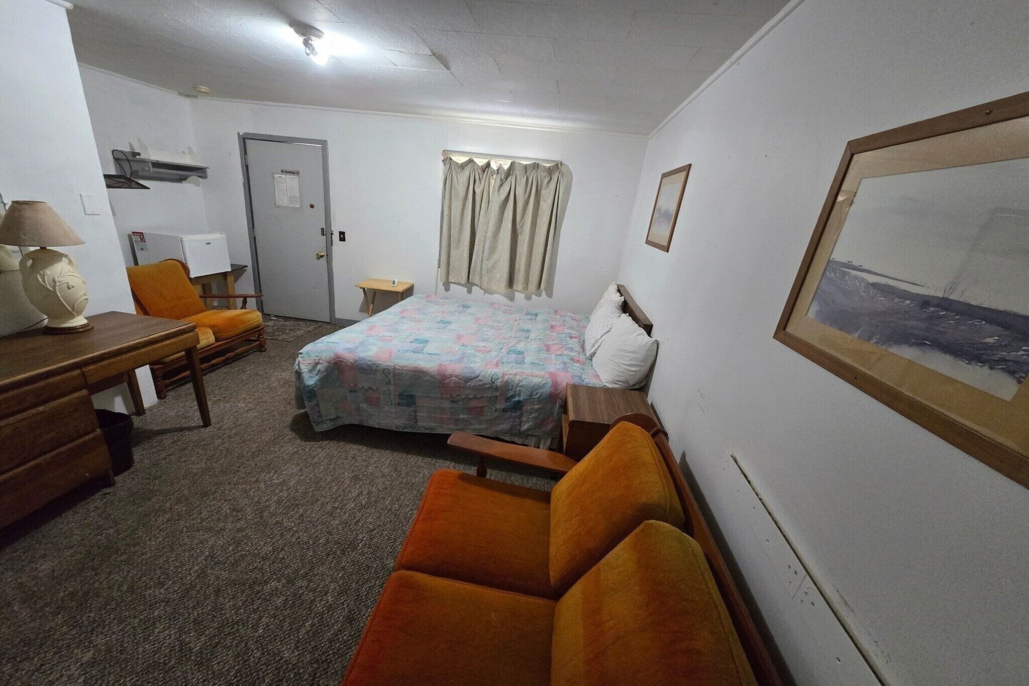 Habitación de hotel con cama, una silla, escritorio, lámpara, un cuadro en la pared y una puerta.