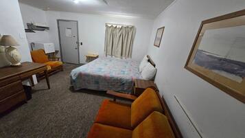 Habitación de hotel con cama, una silla, escritorio, lámpara, un cuadro en la pared y una puerta.