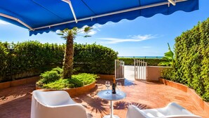 Une terrasse extérieure dotée d’un auvent bleu, de chaises blanches, d’une table avec des verres à vin et offrant une vue sur la mer.