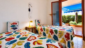 Une chambre avec deux lits, un bureau, une chaise, une lampe et une grande porte-fenêtre coulissante donnant sur un espace extérieur agrémenté de plantes.