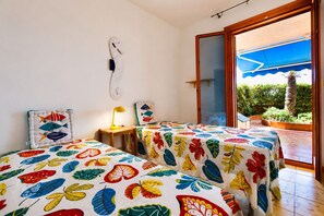 A room with two beds, a desk, a chair, a lamp, and a large sliding glass door leading to an outdoor area with plants. - Vacation Home in Marina Di Ragusa with 3 bedrooms sleeps 6 (Marina Di Ragusa)