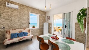 A room with a stone wall, a sofa with blue cushions, a dining table with chairs, and a view of the sea through a window. - La Casa in Riva al Mare by Geco Vacation Rentals (Caucana)