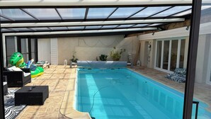A swimming pool with a glass ceiling, surrounded by tiled flooring, wicker furniture, and potted plants. - Chalet between land and sea (Ambrumesnil)