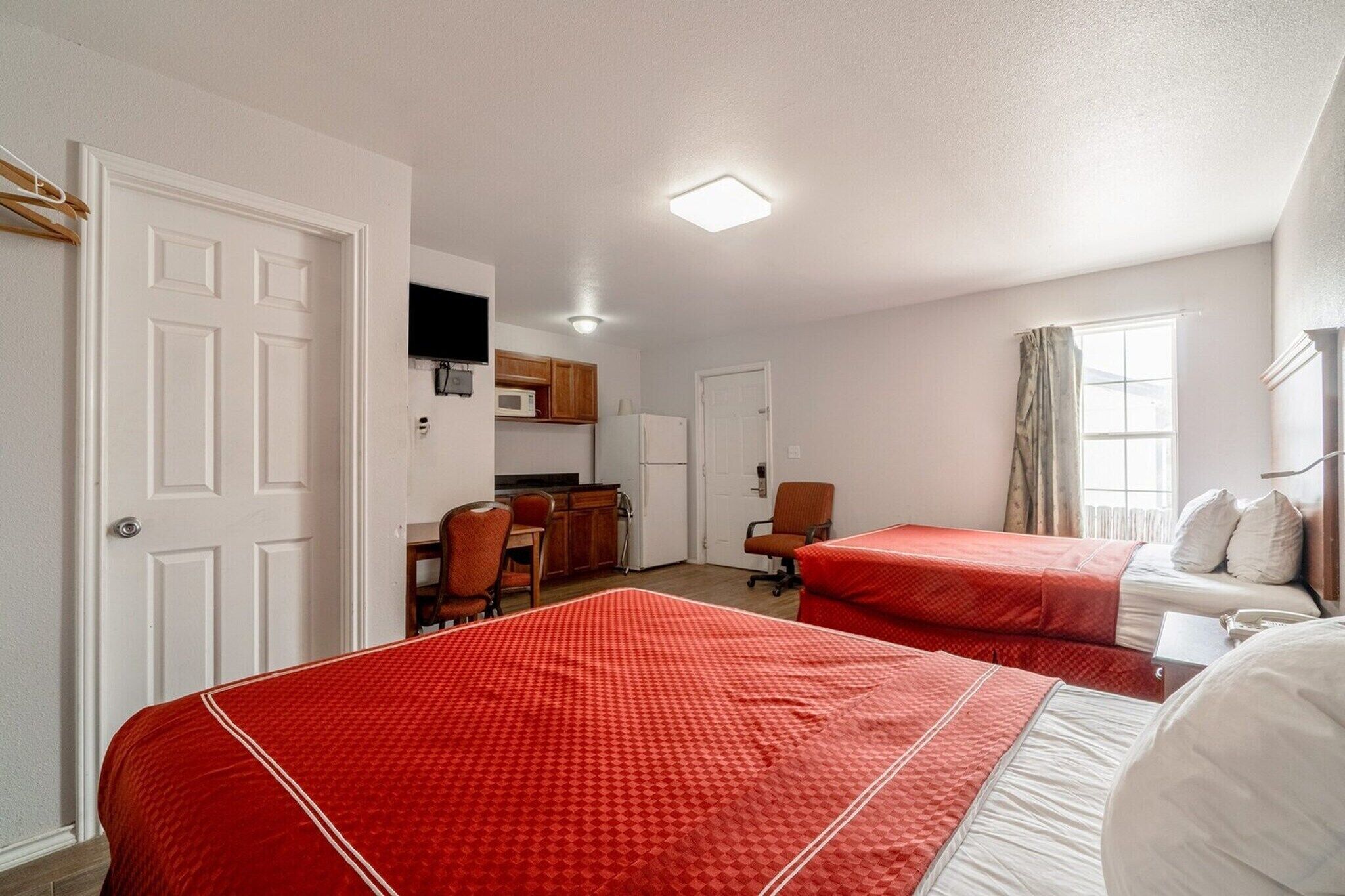 A hotel room with two beds, a kitchenette, a TV, and a window with curtains.