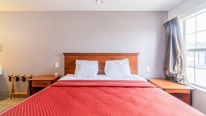 Et hotelværelse med sengegavl i træ, rødt sengetæppe, natbord i træ, skrivebord i træ og et vindue med gardiner.