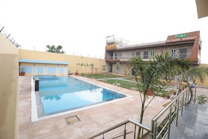 Una piscina con bordeado de baldosas, un edificio con un letrero que dice 'POOL AC ROOMS BANQUET' y un pequeño jardín con plantas en macetas.