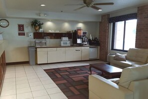 A living room with a sofa, a coffee table, a kitchen area with a microwave, toaster, and coffee machine, and a trash bin.