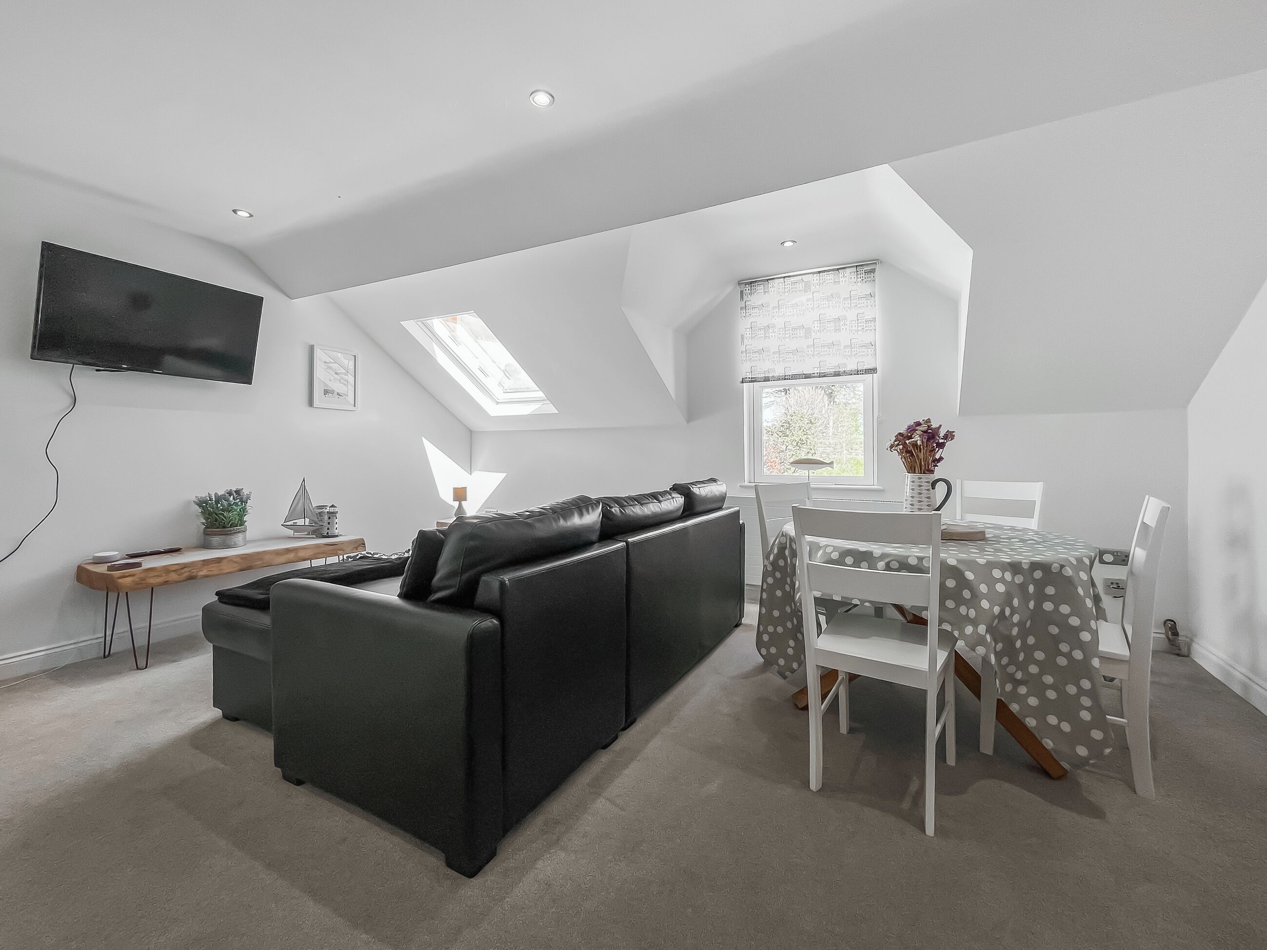 A modern loft apartment with a black leather sofa, a dining table with white chairs, a flat-screen TV mounted on the wall, and skylights.