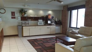A living room with a sofa, a coffee table, a kitchen area with a microwave, toaster, and coffee machine, and a trash bin.