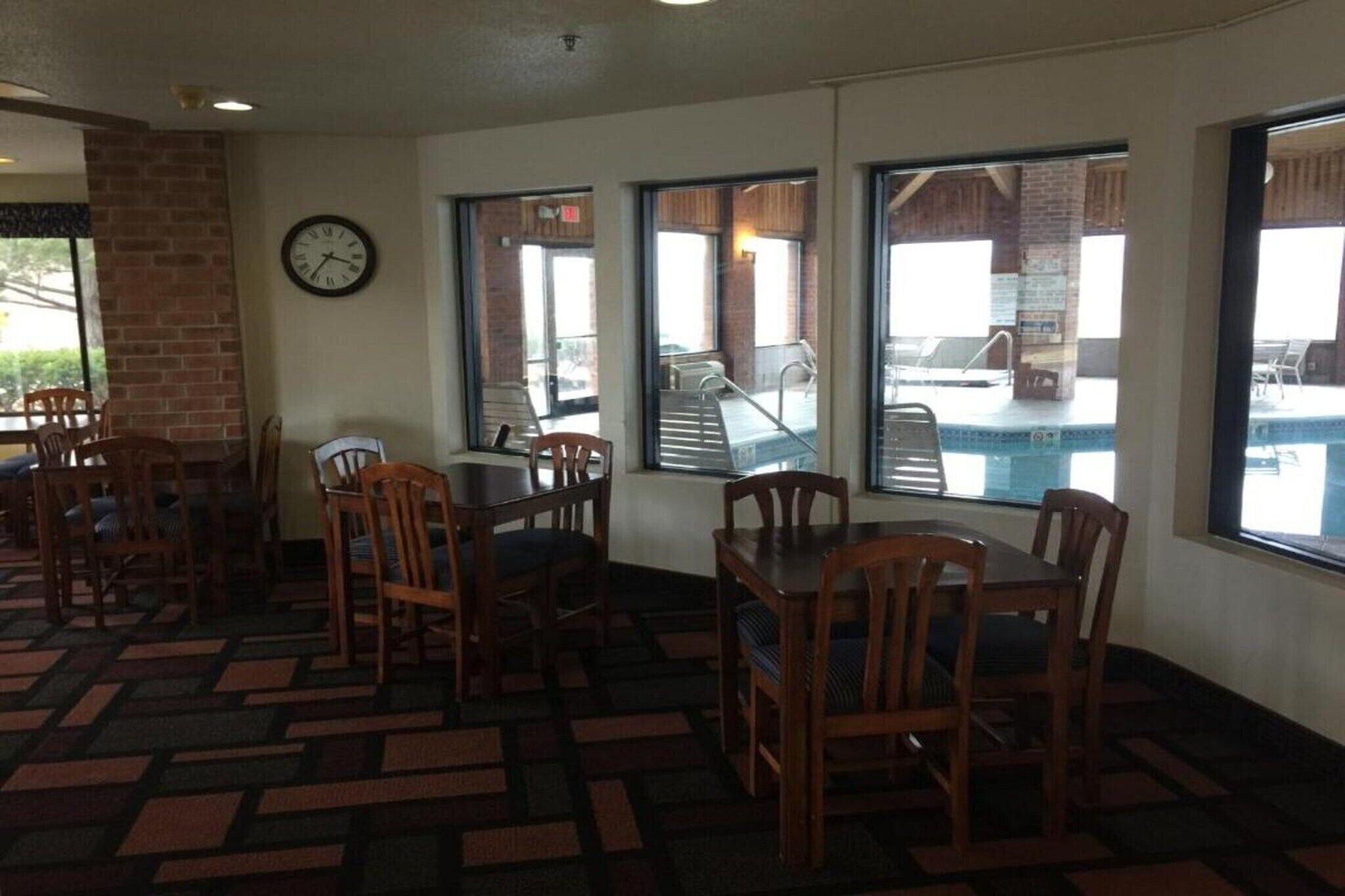 A dining area with wooden tables and chairs, a clock on the wall, and large windows overlooking a pool area.