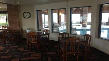 A dining area with wooden tables and chairs, a clock on the wall, and large windows overlooking a pool area.