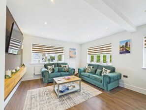 A spacious living room with two green sofas, a coffee table, and a large window with blinds. - 4 bedroom accommodation in Whitstable (Whitstable)