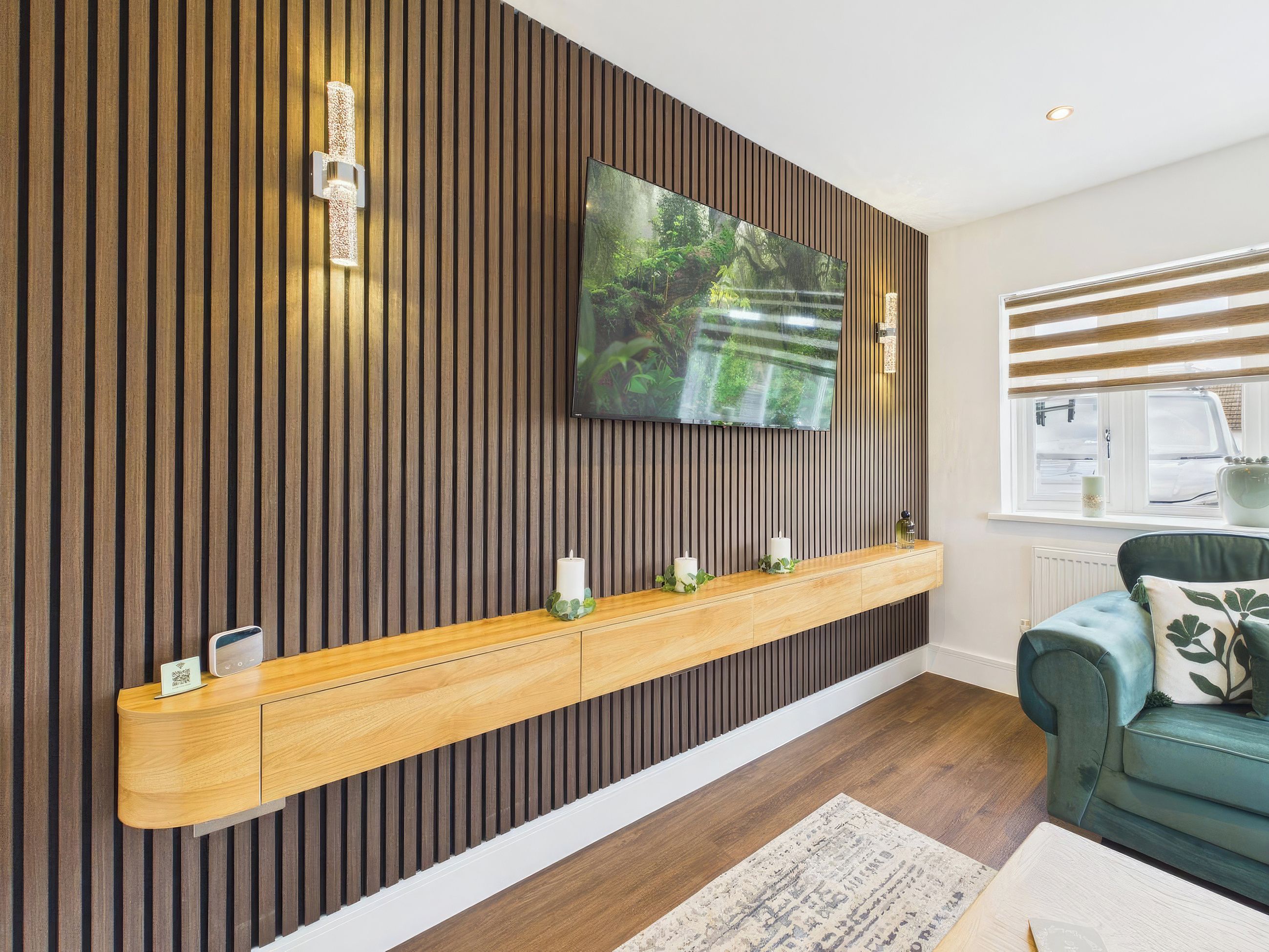 A modern living room with a wooden slatted wall, a mounted TV, a wooden console table with candles, a green sofa, and a window with blinds.
