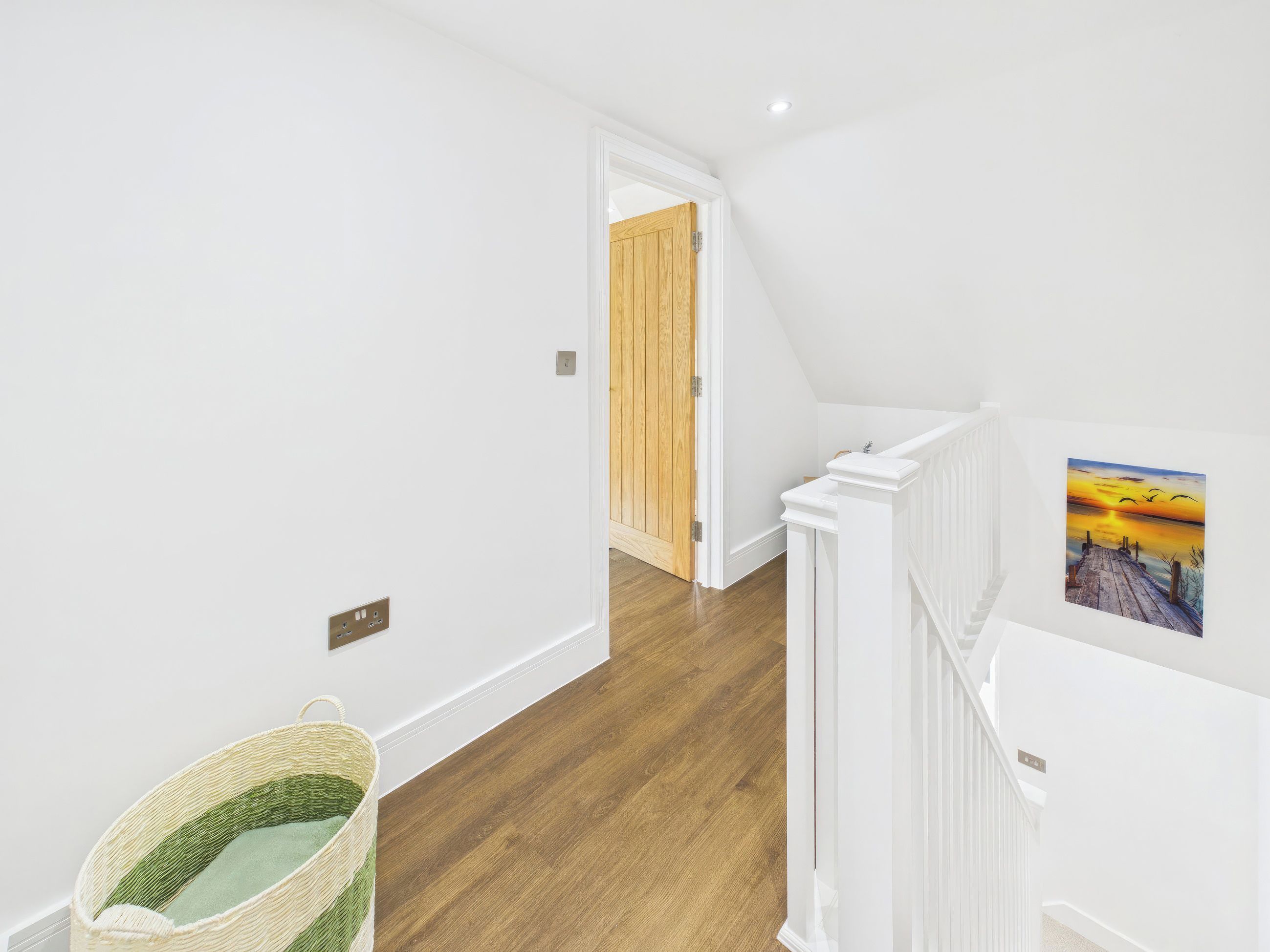 A hallway with wooden flooring, white walls, a wooden door, and a white railing with a picture.
