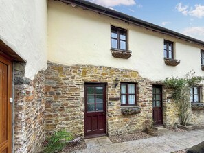 A stone facade with a cream-colored upper wall, a wooden door, and a window with a hanging basket. - Brambles Cottage - UK51981 (Little Torrington)