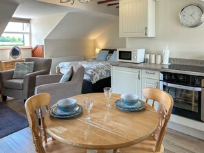 Ein gemütliches Zimmer mit einem Bett, einem Esstisch mit Stühlen, einer Mikrowelle, einem Wasserkocher und einer Wanduhr.
