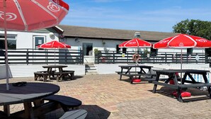 An outdoor patio area with picnic tables and red umbrellas, a white building with a sloped roof, and a fenced area. - 3 steps to entrance. (Rhydlewis)